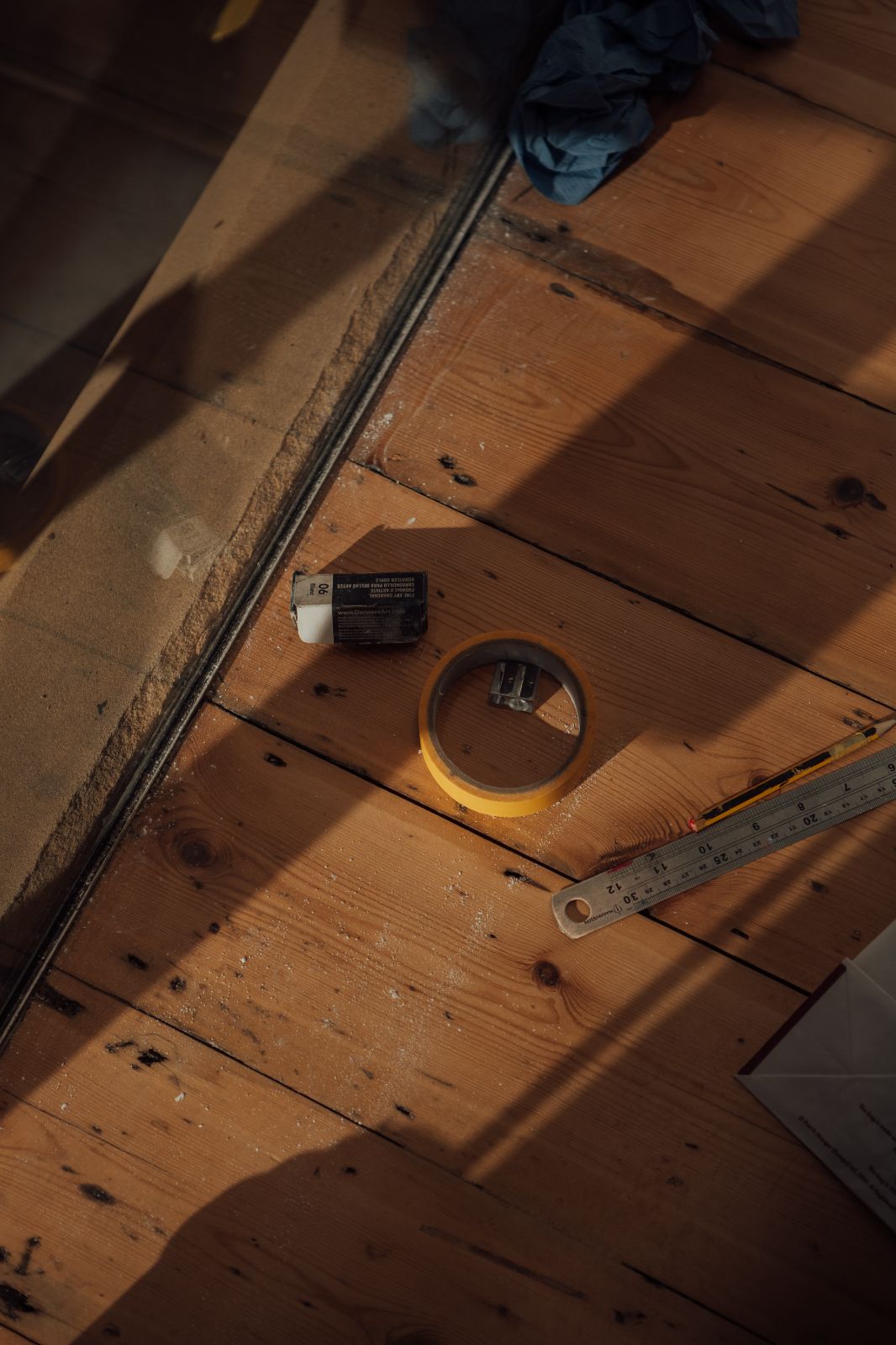 Tape, pencil sharpener, pencil and ruler on wooden floorboards with strong shadow cast by unseen object.