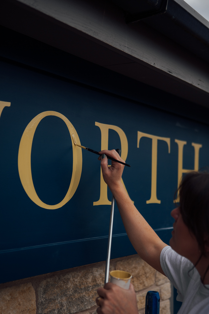 Signwriter Mia Warner painting a gold O on the blue facia sign of Chatsworth Kitchen’s sign at Peak Village. 