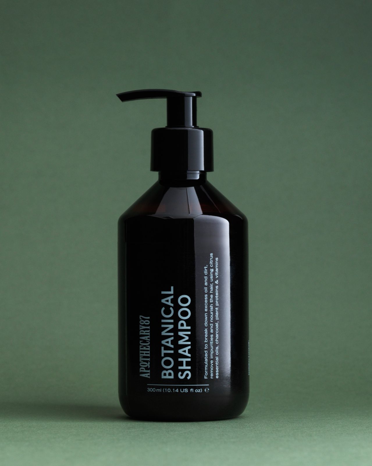 A brown pump bottle of Apothecary87 BOTANICAL SHAMPOO on green warmly-lit studio background.