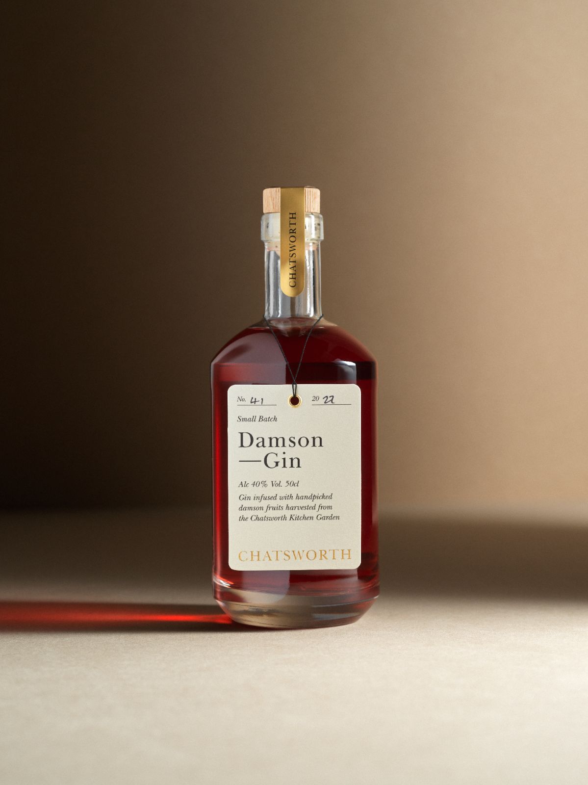 A bottle of red small batch Chatsworth Damson Gin. Clear glass with a cork stopper, gold sticker and cream label.