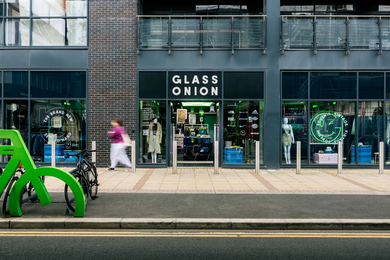 Outside of Glass Onion’s clothing store with a green winking smiley face light in window. Green bike rack in foreground