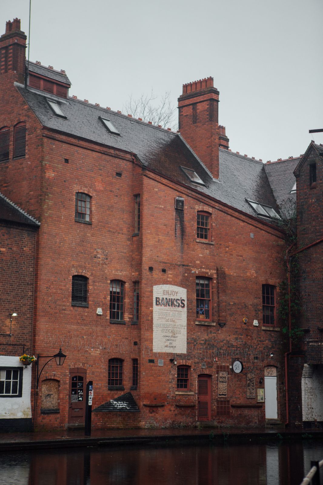 Tall Victorian red-brick building, with painted sign advertising ales, along a Birmingham canal on a rainy winter’s day.