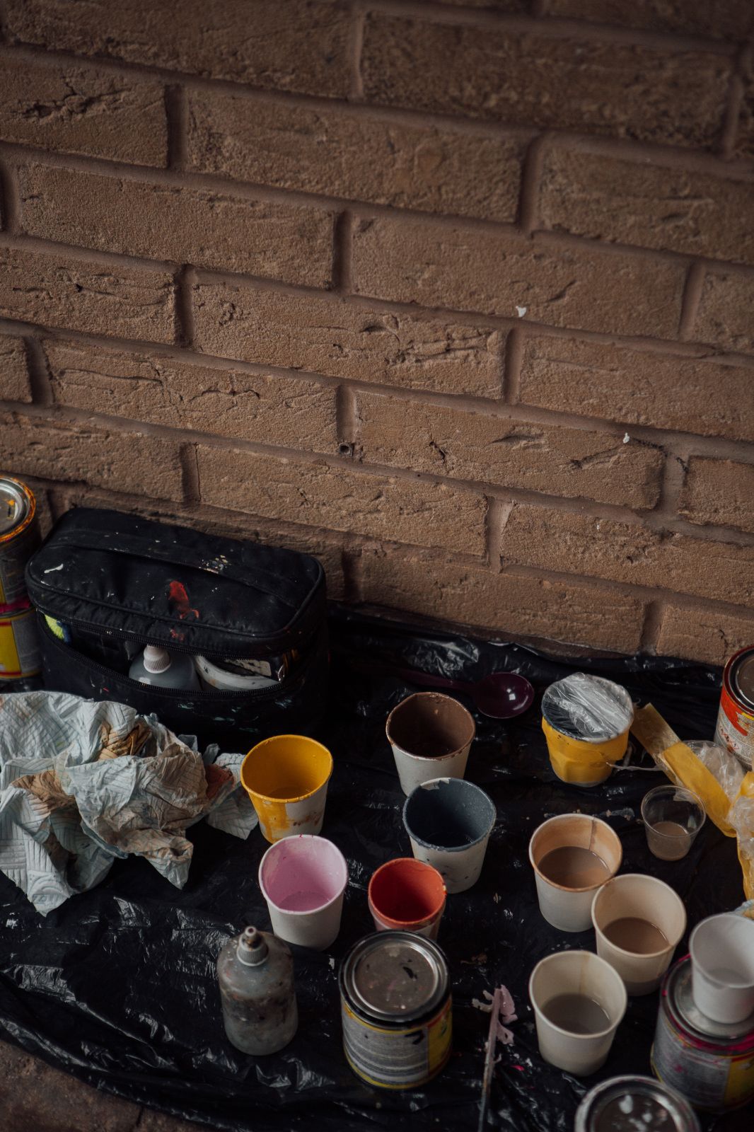 Paper cups of paint in different colours, rags and paint tins on black plastic sheeting on the ground along a brick wall. 