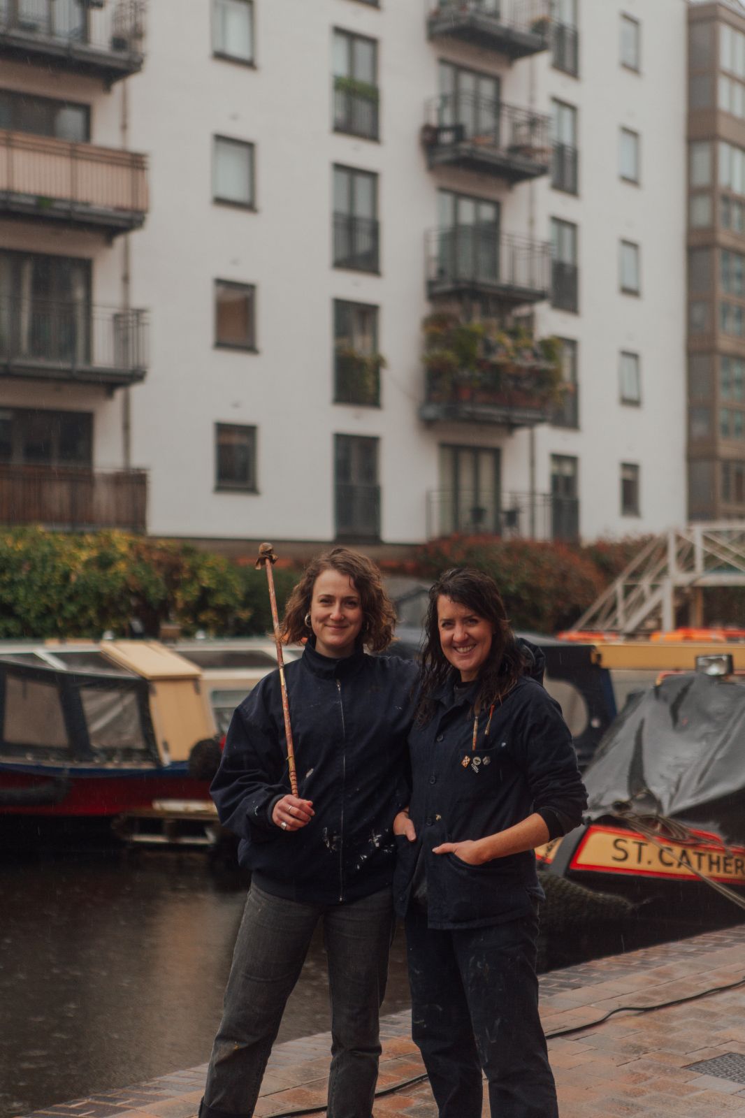 Two female signwriters in dark work clothing, one holds a mahl stick. They sand in front of a wharf with narrow boats.