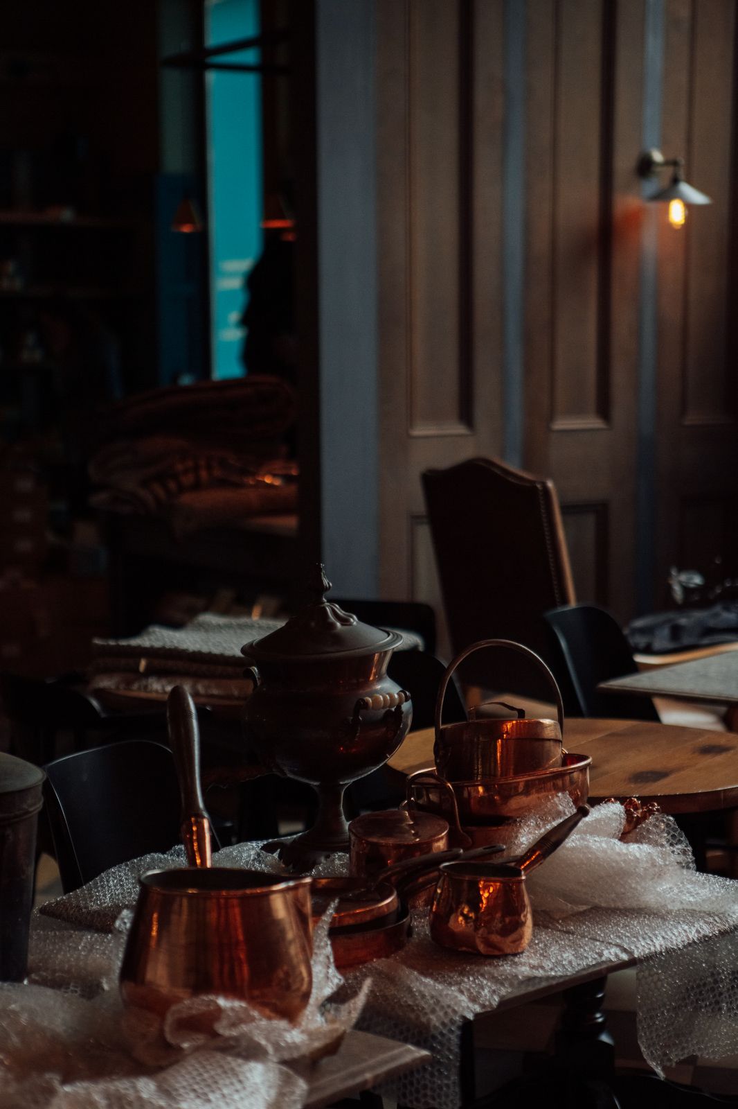 Antique copper pots and pans stacked on a couple tables surrounded by bubble wrap with wood panelling in the background.