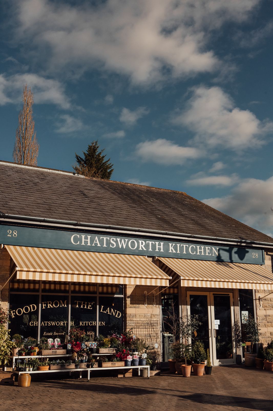 External view of Chatsworth Kitchen.  Blue facia with yellow and white striped awning. Warm light, blue sky with clouds