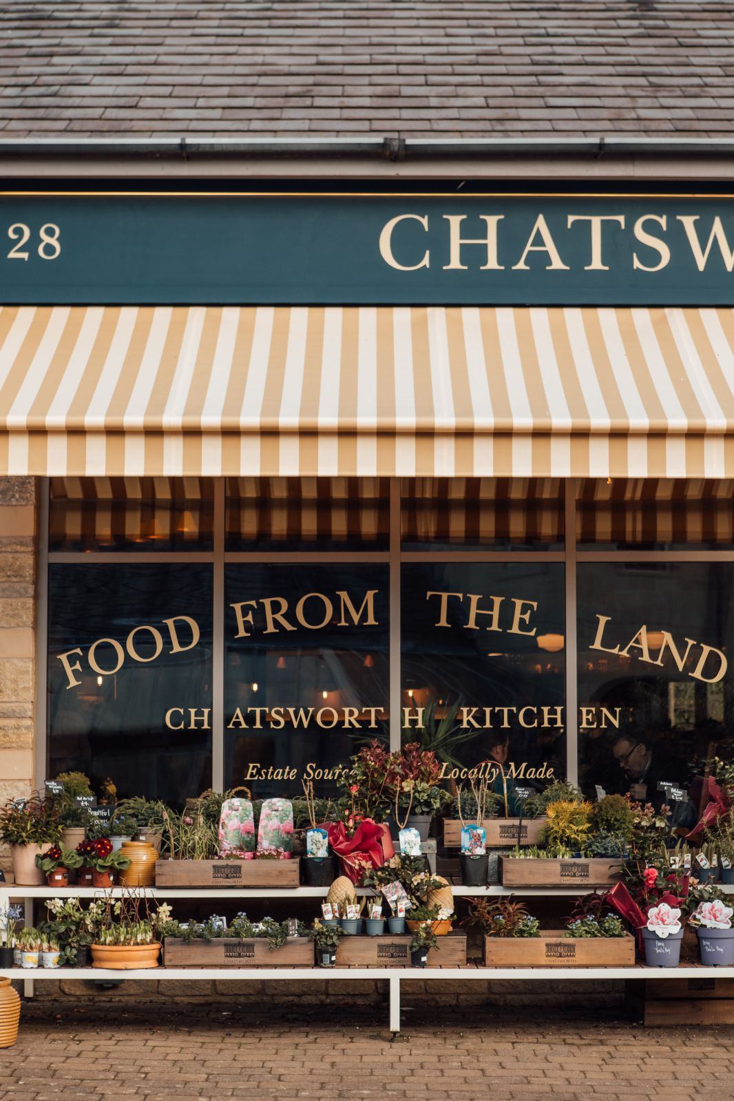 Chatsworth Kitchen facia. Sign writing on windows FOOD FROM THE LAND. Pots of flowers and shrubs outside the building.