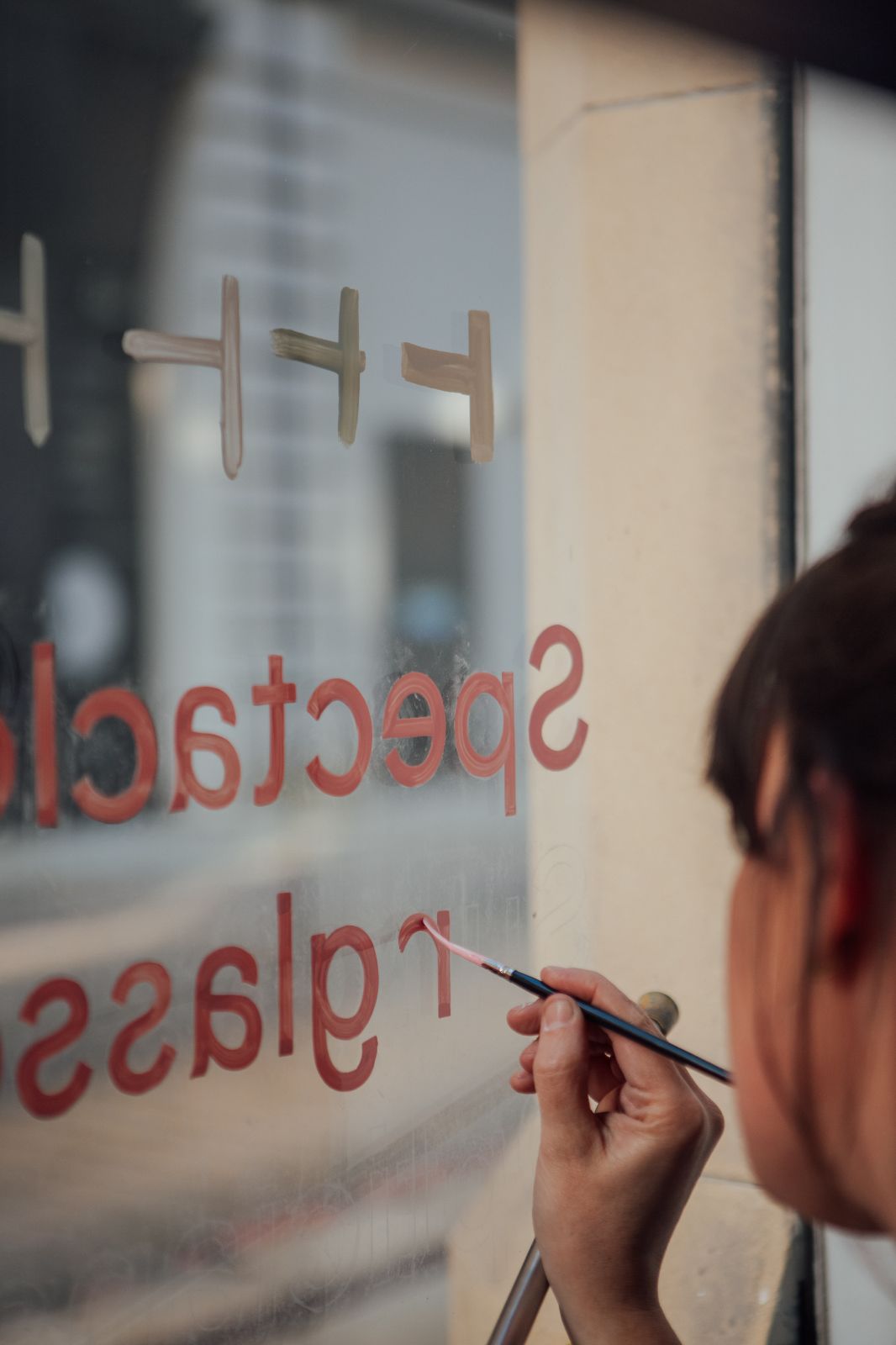 Signwriter Mia Warner painting pink letters on a glass window shop front. Chalk stencil lines for the words are visible.