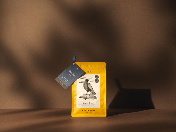 Dark woods coffee - Yellow Crow Tree packaging, with branding by 93 on dark brown background - warm studio lighting