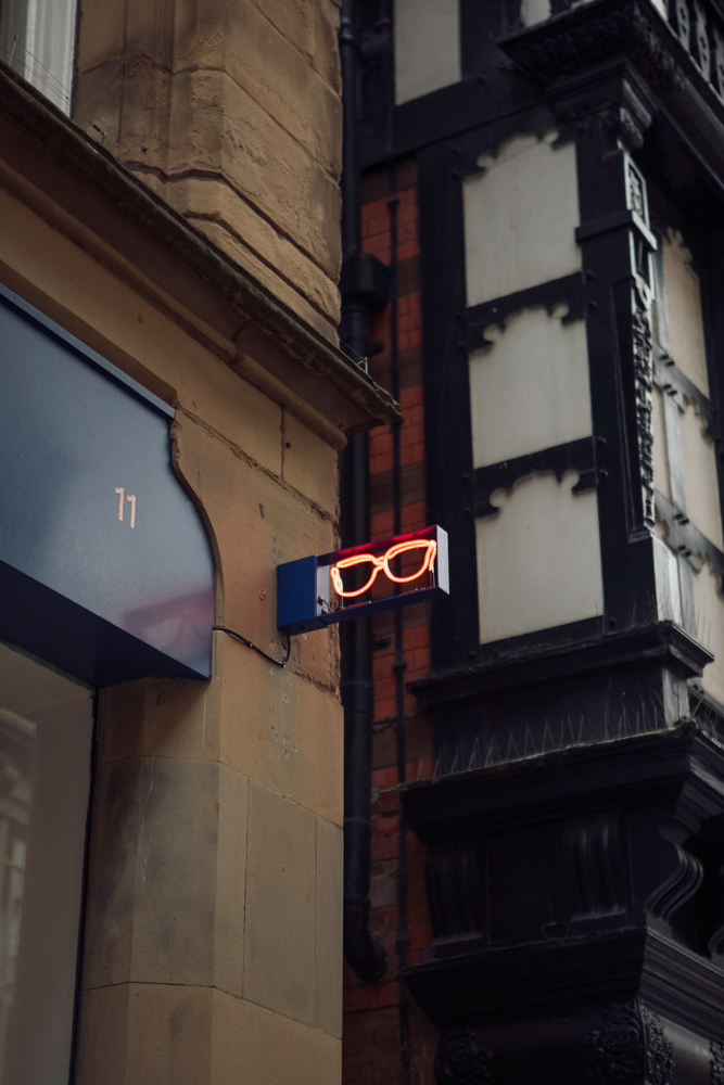 Corner of building with small overhanging pink neon sign of a pair of glasses on blue backing & old buildings in background