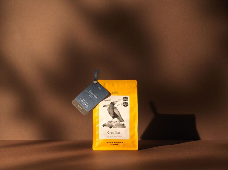 Darkwoods coffee - Yellow Crow Tree packaging, with branding by 93 on dark brown background - warm studio lighting