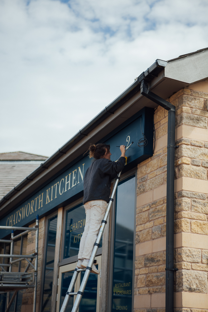 Signwriter Mia Warner on a ladder painting a 2 in gold on the blue facia above the entrance of Chatsworth Kitchen farm shop. 