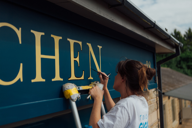 Signwriter painting a black outline around the gold letters of Kitchen on the dark blue shop front facia. 