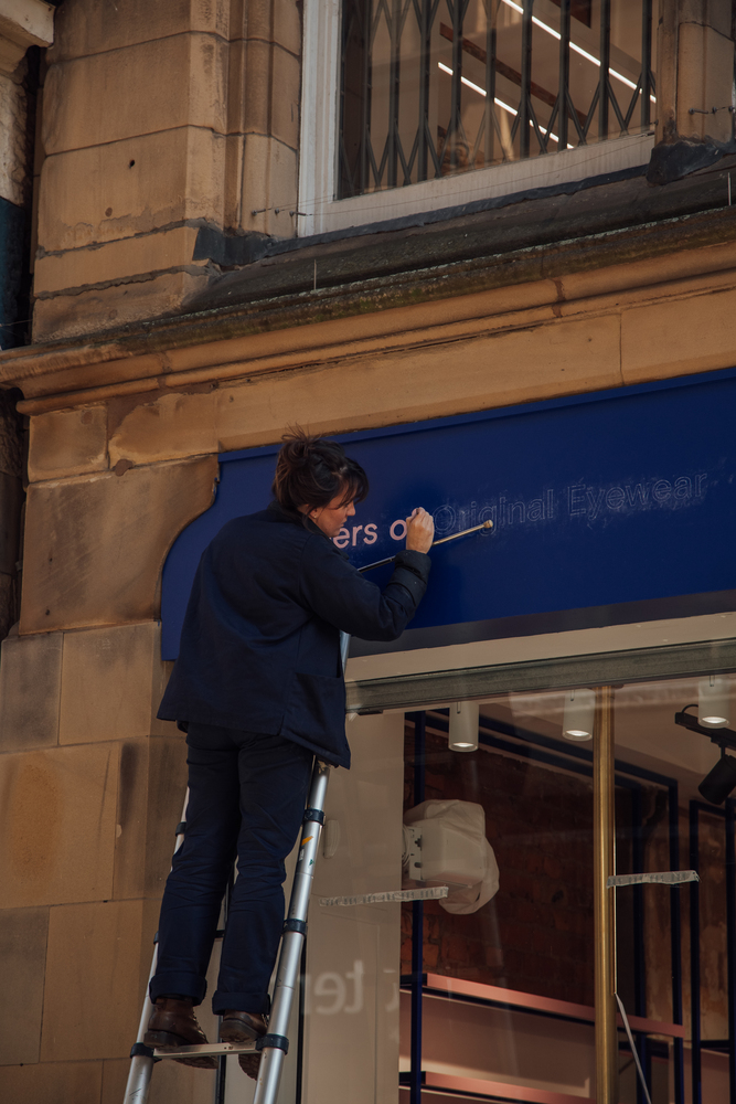 Signwriter on a ladder painting pink letters on a royal blue shop facia of an elegant stone building in Manchester