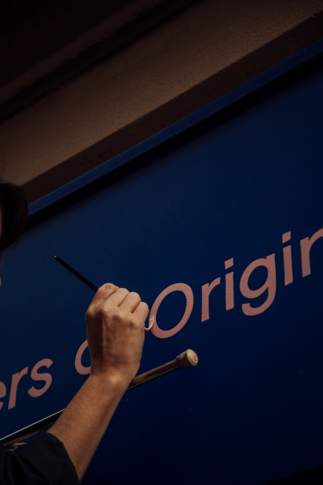 Signwriter painting the word Original in pink letters with a fine brush on a blue shop facia. The hand rests on a mahlstick 