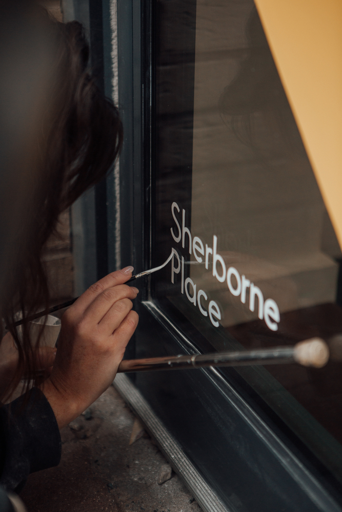 Signwriter Mia Warner painting the words Sherborne Place in white on a glass door. Her hand rests on a mahlstick