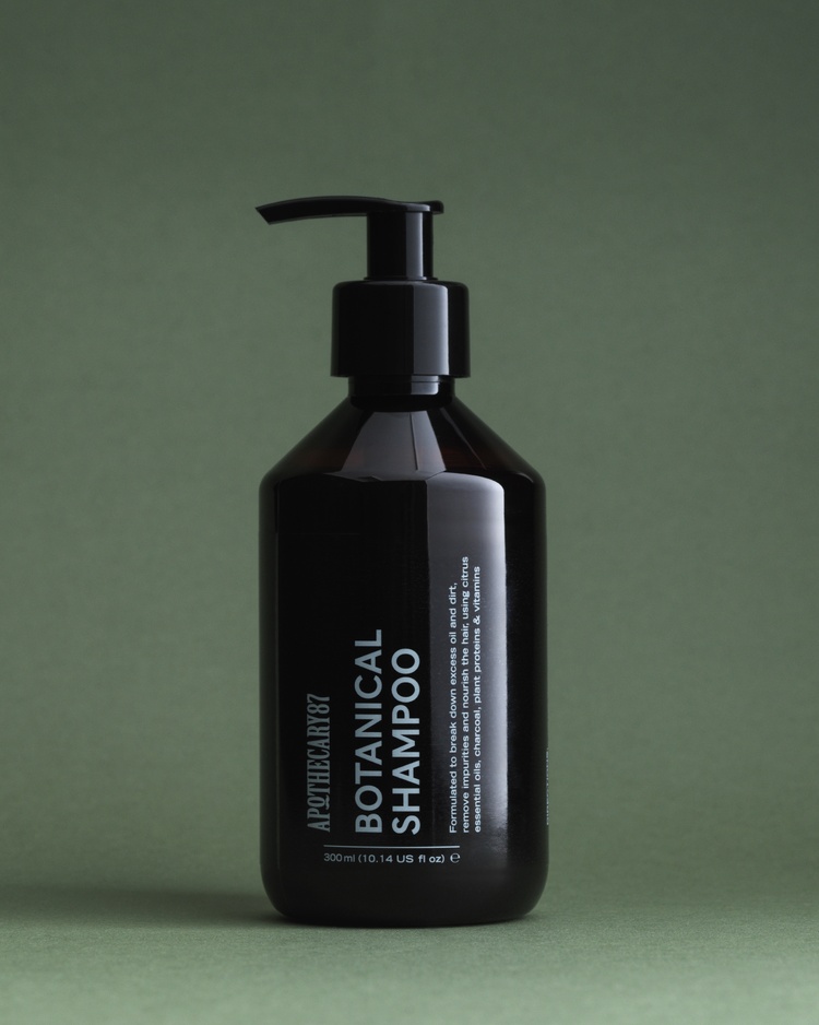 A brown bottle of Apothecary 87 Botanical Shampoo on a green studio background. Packaging design by 93ft.