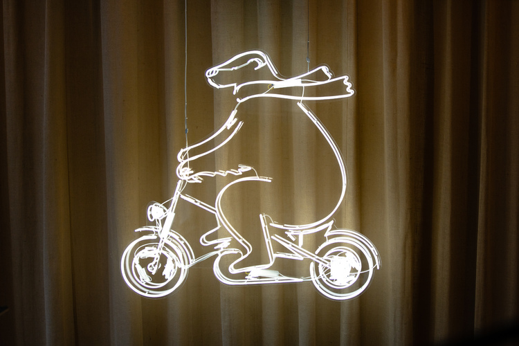 White glass neon sign of a simple illustrated bear wearing a scarf and riding a bicycle, hung in front of linen curtains