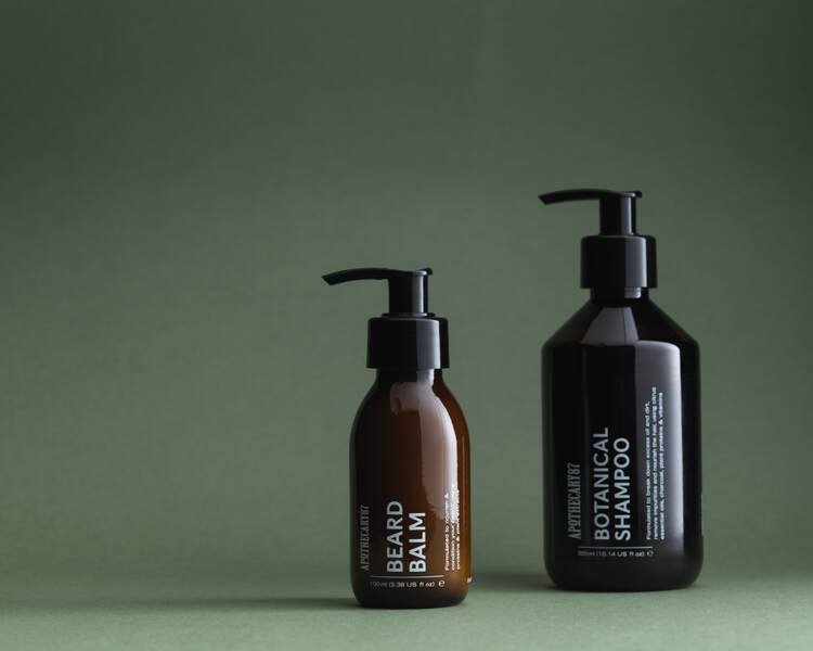 4 brown glass bottles of Apothecary87 salt tonic, beard oil, beard balm, botanical shampoo. Warm studio lighting.