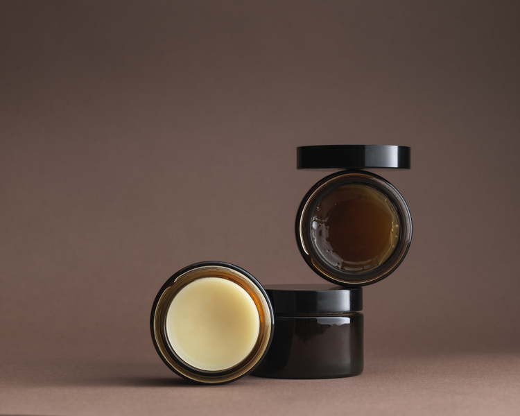 3 brown glass pots stacked artistically with contents facing forward - shiny amber gel and cream pomade. Lid balanced on top.