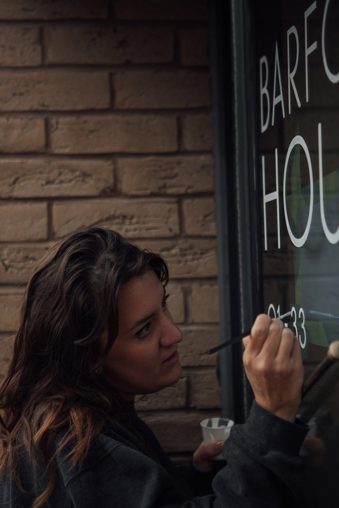 Female signwriter with brown hair painting 33 in white on a glass door. She tilts her head slightly and hand rests on a mahl. 