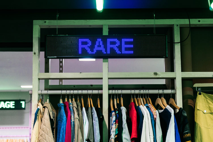 Digital L.E.D signage spelling Rare in purple above a rail of vintage clothing hung from a plain timber frame. 