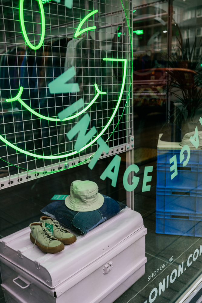 L.E.D green tube light in the shape of a smiley face & pink chest displaying shoes, bucket hat & denim cushion in shop window