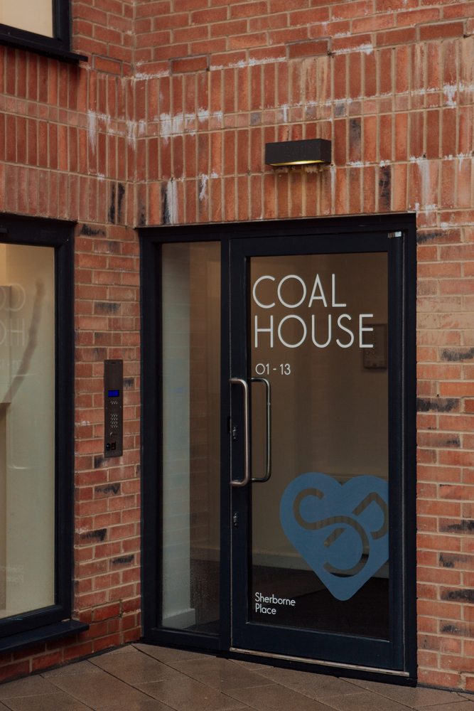 New red brick building with black-framed glass door. Painted on the glass is COAL HOUSE 01-13 and a heart logo with an S.P.