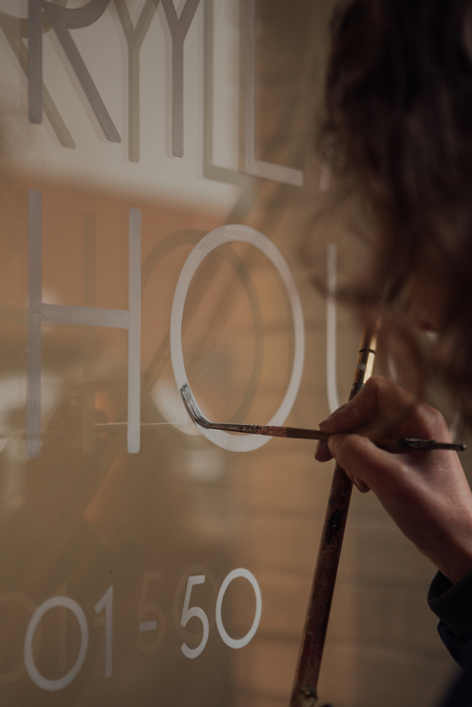 Signwriter painting a white letter O on a glass door while her hand rests on a mahl stick.