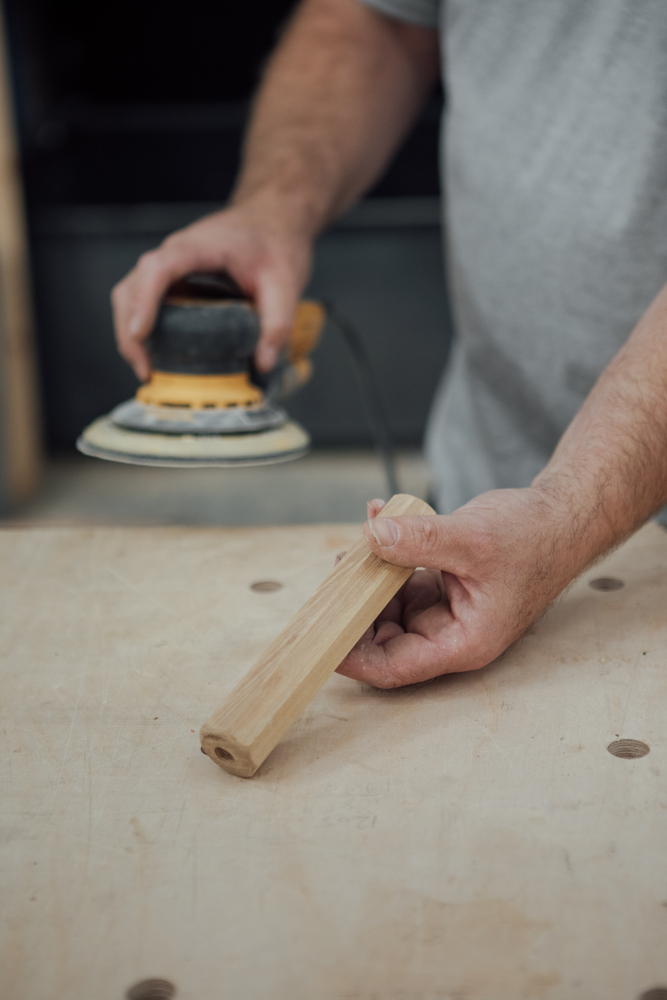 Close up of man holding an electric sander in one hand & octagonal turned wooden handle in the other on a wooden work bench. 