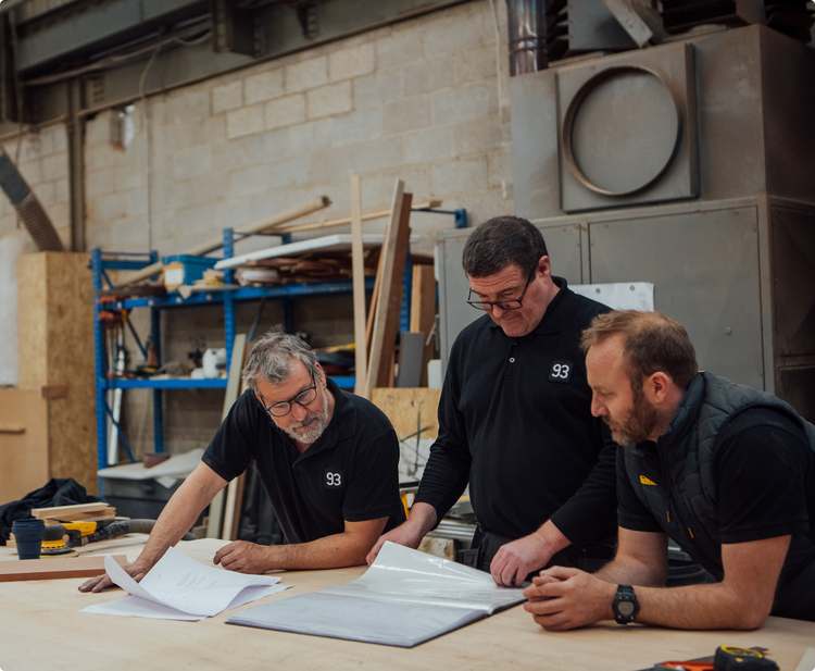 Three men in black polo shirts looking at paper plans in 93 Makes bespoke furniture & fittings workshop in Sheffield. 