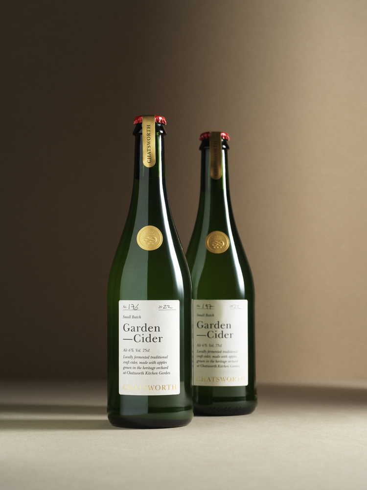 Two green bottles of Garden Cider with a red beer cap and gold embossed sticker. Products shot in a warmly lit studio.